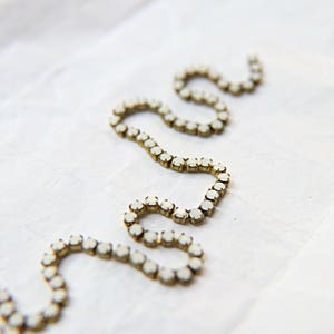 Puede incluir: Primer plano de una cadena de joyer&iacute;a decorativa con peque&ntilde;as piedras preciosas blancas y redondas engastadas en metal dorado. La cadena est&aacute; dispuesta en un patr&oacute;n ondulado sobre un fondo blanco texturizado.