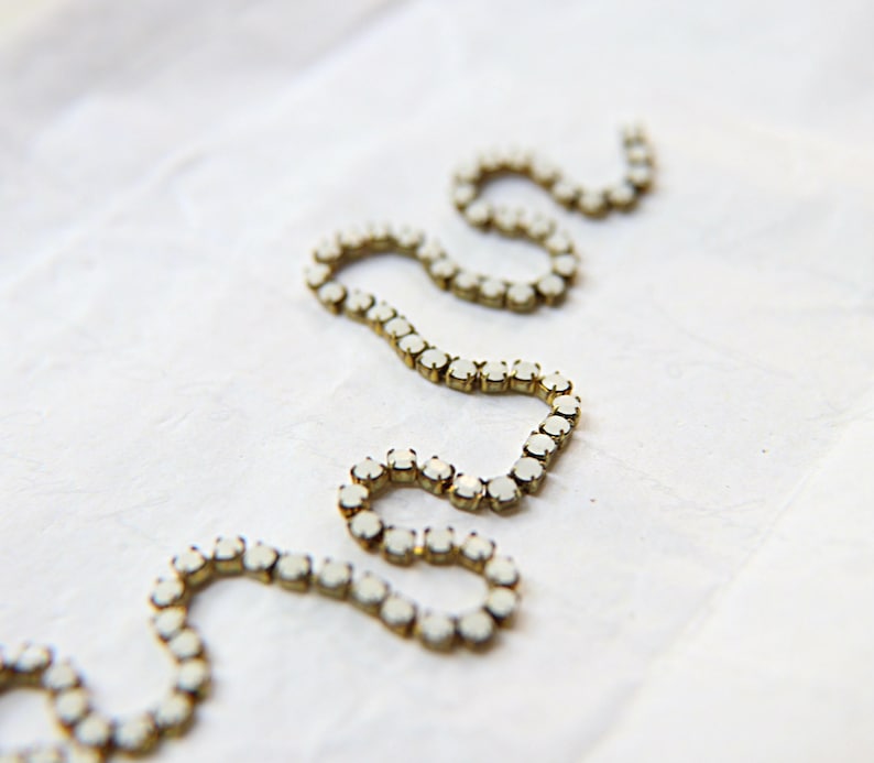 Puede incluir: Primer plano de una cadena de joyer&iacute;a decorativa con peque&ntilde;as piedras preciosas blancas y redondas engastadas en metal dorado. La cadena est&aacute; dispuesta en un patr&oacute;n ondulado sobre un fondo blanco texturizado.