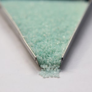 May include: A close-up of a metal tray filled with light blue glass beads. The beads are spilling out of the tray onto a white surface.
