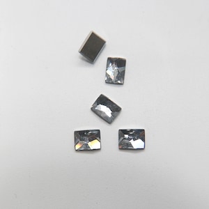 May include: Five clear glass rectangular rhinestones on a white background. The rhinestones are different sizes and are arranged in a random pattern.