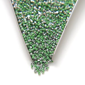 May include: A close-up of a metal container filled with green and silver seed beads. The beads are spilling out of the container onto a white surface.
