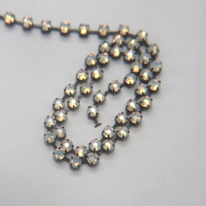 Pode incluir: Um colar delicado com pedras preciosas redondas e iridescentes, engastadas numa corrente de metal escuro. As pedras preciosas têm uma qualidade subtil e brilhante, refletindo a luz com um brilho opalescente suave.