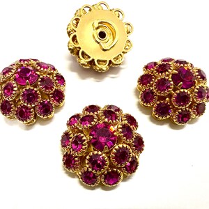 May include: Four round buttons featuring a gold-tone metal base encrusted with multiple faceted fuchsia pink rhinestones. The buttons have a domed shape with a central stone surrounded by smaller stones, and one button shows the metal shank on the back.