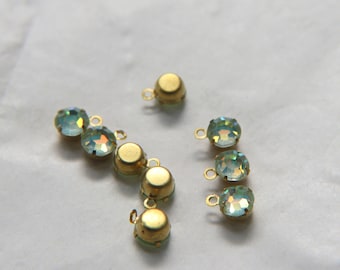 Puede incluir: Primer plano de varios pequeños dijes de metal dorado con cristales iridiscentes, redondos, de color verde claro y amarillo. Cada dije tiene un pequeño lazo en la parte superior. Los dijes están dispersos sobre una superficie blanca.