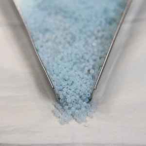May include: A metal scoop filled with light blue seed beads. The beads are spilling out of the scoop onto a white surface.