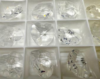 Puede incluir: Una colección de cuentas de cristal transparente facetadas en varias formas, incluyendo corazones y lágrimas, se disponen en una bandeja blanca dividida. Los cristales reflejan la luz, creando un efecto brillante. La bandeja está dividida en múltiples compartimentos.