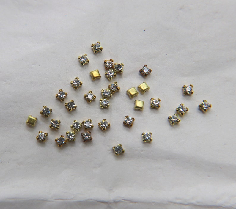 Peut inclure: Une collection de petits strass transparents et de d&eacute;corations en m&eacute;tal dor&eacute;. Les strass sont de forme carr&eacute;e et ont un design facett&eacute;. Les d&eacute;corations en m&eacute;tal sont &eacute;galement de forme carr&eacute;e et ont une finition lisse.