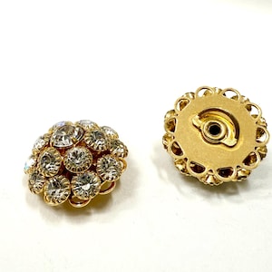 Può includere: Due orecchini a clip vintage color oro. Ogni orecchino presenta un gruppo di strass rotondi e trasparenti incastonati in metallo color oro. Il retro di un orecchino è visibile, mostrando il meccanismo a clip e un bordo decorativo.