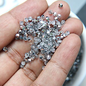 Peut inclure: Une main tenant une grande quantité de perles de verre de couleur argent. Les perles sont facettées et ont une finition brillante.