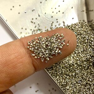May include: A collection of tiny, faceted, metallic-colored stones with a pointed, conical shape. These small, reflective gems are scattered across a silver-toned tray and are also gathered in a small pile on a finger for display.