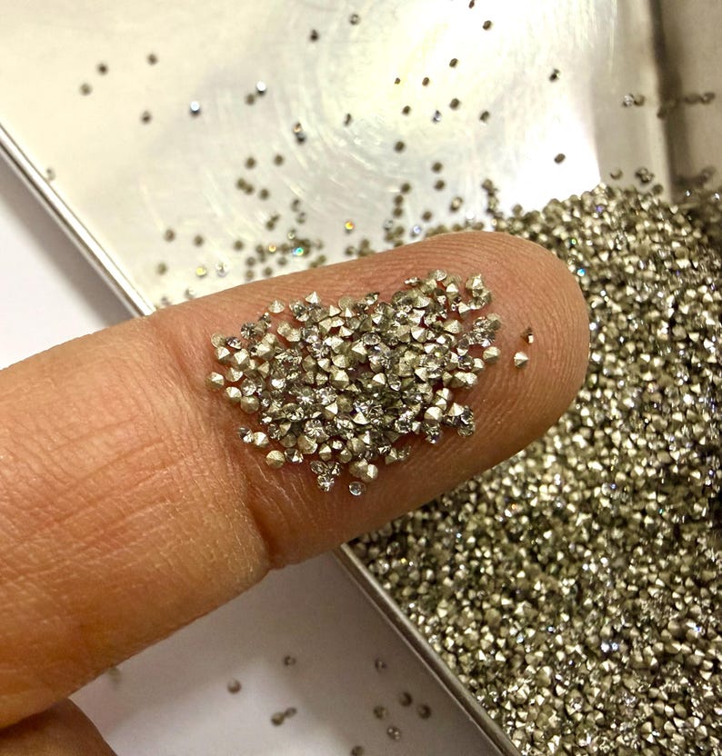 May include: A collection of tiny, faceted, metallic-colored stones with a pointed, conical shape. These small, reflective gems are scattered across a silver-toned tray and are also gathered in a small pile on a finger for display.