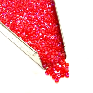 Pode incluir: Uma colher de metal cheia de pequenas contas vermelhas vibrantes. As contas estão a cair sobre uma superfície branca. As contas são uma mistura de vermelho sólido e translúcido, criando um efeito brilhante. Ideal para fazer joias.