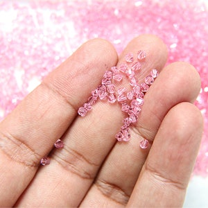 May include: Close-up of small, faceted, light pink beads. The beads are scattered across a hand, with a blurred background of more beads. These beads are likely used for jewelry making or crafting.