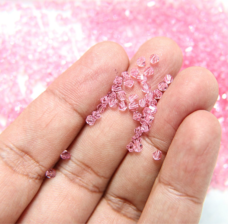 May include: Close-up of small, faceted, light pink beads. The beads are scattered across a hand, with a blurred background of more beads. These beads are likely used for jewelry making or crafting.