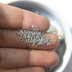 May include: A collection of tiny, multi-colored craft glitter particles in shades of silver, teal, and soft pink. These small, reflective, and granular pieces are loose and scattered, showcasing a metallic finish and a faceted texture.