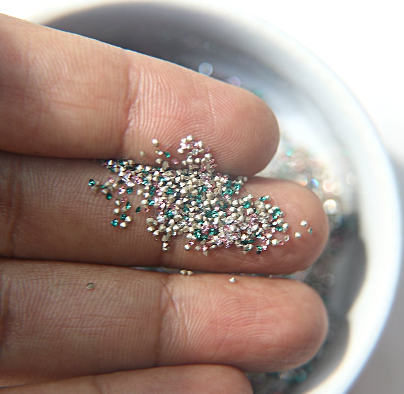 May include: A collection of tiny, multi-colored craft glitter particles in shades of silver, teal, and soft pink. These small, reflective, and granular pieces are loose and scattered, showcasing a metallic finish and a faceted texture.