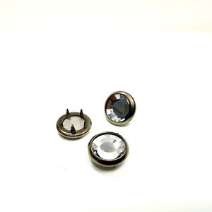 May include: Three silver-toned snap fasteners with clear, faceted crystal centres. One snap is disassembled, showing the pronged base. The other two snaps are assembled, displaying the sparkling crystals. These are ideal for adding a decorative touch to clothing or accessories.