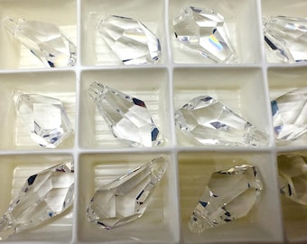 Puede incluir: Primer plano de colgantes de cristal transparente y facetado dispuestos en una bandeja blanca dividida. Los cristales tienen múltiples facetas que reflejan la luz, creando un efecto brillante. Los colgantes tienen forma de lágrima y rectangular.