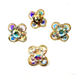 Può includere: Quattro orecchini vintage a forma di fiore con cristalli iridescenti e montature in metallo dorato. Ogni orecchino presenta un cristallo centrale circondato da cristalli più piccoli e colorati, creando un design floreale scintillante. Gli orecchini sono su sfondo bianco.