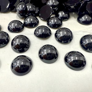 May include: A collection of glossy, black, half-dome cabochons. These smooth, rounded stones reflect light, creating a mirror-like surface. The cabochons are scattered, with some overlapping, suggesting they are loose components for jewelry or crafts.