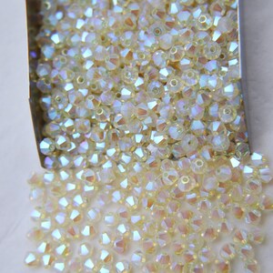 May include: A close-up of a container filled with iridescent, light yellow, faceted glass beads. The beads are spilling out of the container onto a white surface.