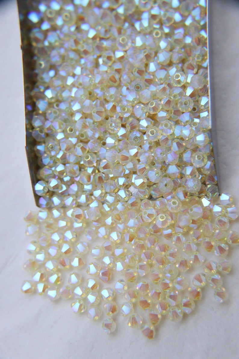 May include: A close-up of a container filled with iridescent, light yellow, faceted glass beads. The beads are spilling out of the container onto a white surface.
