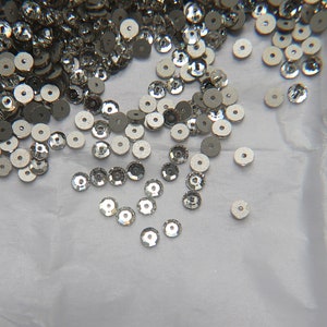 May include: A pile of silver-colored sequins scattered on a white surface. The sequins are round and have a slightly raised center.