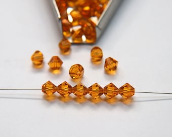 Puede incluir: Una hebra de cuentas de vidrio de color ámbar enhebradas en un hilo fino. Las cuentas son facetadas y tienen una forma de diamante. También hay varias cuentas sueltas esparcidas alrededor de la hebra.