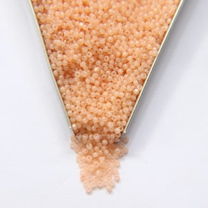 May include: A close-up of a container filled with small, peach-colored beads. The beads are spilling out of the container onto a white surface.