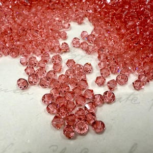 Könnte beinhalten: Eine Ansammlung facettierter, rosafarbener Perlen. Die Perlen sind auf einer weißen Oberfläche verstreut, wobei ein Teil dicht gepackt ist. Die Perlen haben ein funkelndes, kristallines Aussehen und reflektieren das Licht.