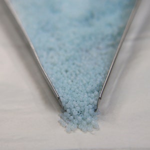 May include: A metal scoop filled with light blue seed beads, spilling out onto a white surface.