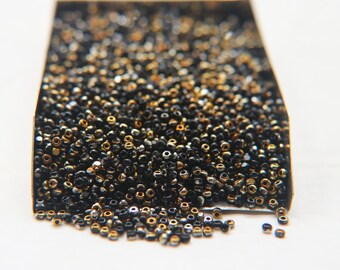 May include: A close-up of a box filled with black and gold seed beads. The beads are spilling out of the box onto a white surface.