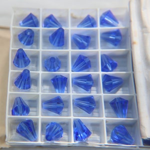 Op de afbeelding: Een doorzichtige plastic container gevuld met 24 blauwe kristallen kralen in de vorm van een kegel. De kralen zijn gerangschikt in een rasterpatroon.