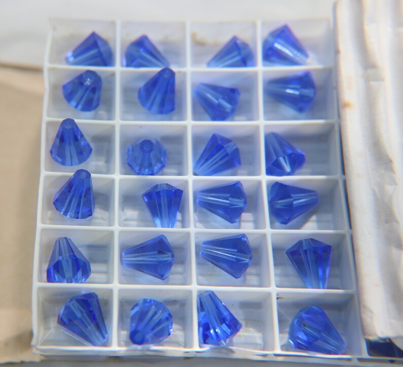 Op de afbeelding: Een doorzichtige plastic container gevuld met 24 blauwe kristallen kralen in de vorm van een kegel. De kralen zijn gerangschikt in een rasterpatroon.