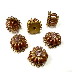 Può includere: Sei pomelli per cassetti in metallo color rame in stile vintage con un design floreale. Ogni pomello presenta un cristallo rosa centrale circondato da cristalli trasparenti più piccoli. I pomelli hanno un design traforato decorativo e sono su sfondo bianco.