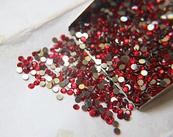 May include: A close-up image of a small metal scoop filled with red rhinestones. The rhinestones are spilling out of the scoop onto a white surface.