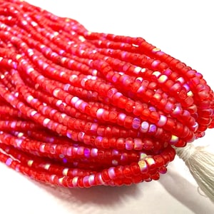 May include: A collection of bright red seed beads with iridescent highlights. The beads are strung together, creating multiple strands. A white tassel is visible at the end. Suitable for jewellery making and crafts.