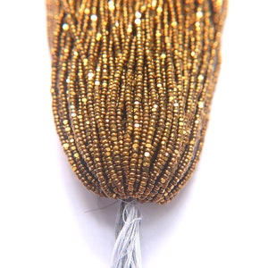 Puede incluir: Primer plano de un paquete de cuentas de semillas doradas. Las cuentas son peque&ntilde;as y tienen un ligero brillo met&aacute;lico.