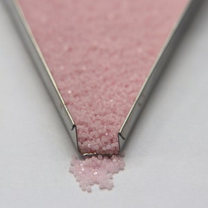 May include: A close-up of a metal tray filled with pink seed beads. The beads are spilling out of the tray onto a white surface.