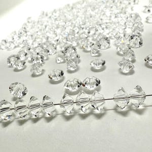 May include: A collection of clear crystal beads, some strung on a wire, are displayed on a white surface. The beads vary in shape and size, with a faceted design that catches the light, creating a sparkling effect.