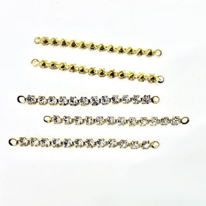 Può includere: Diversi connettori a catena di strass in tonalità oro e argento sono esposti su sfondo bianco. I connettori sono dritti, con piccoli anelli ad ogni estremità. Gli strass trasparenti sono ravvicinati.