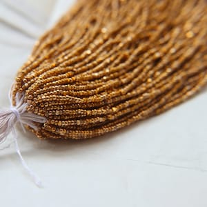 May include: A close-up of a bundle of gold-coloured seed beads. The beads are small and have a slightly metallic sheen.