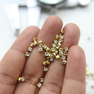 Peut inclure: Une main tenant un tas de petits strass carr&eacute;s dor&eacute;s avec des cristaux transparents au centre.