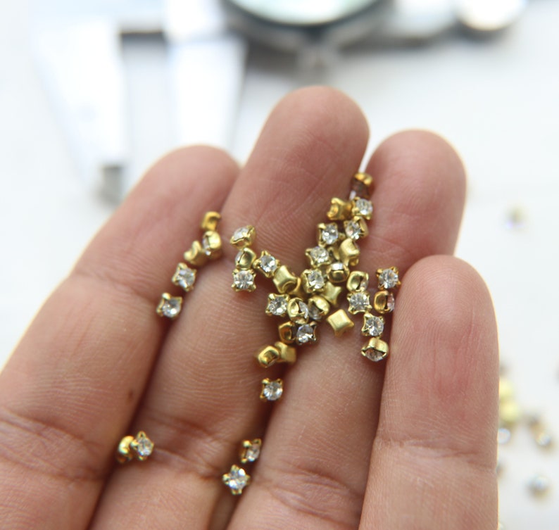 Peut inclure: Une main tenant un tas de petits strass carr&eacute;s dor&eacute;s avec des cristaux transparents au centre.