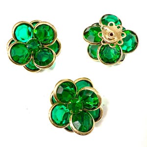 Può includere: Tre orecchini a forma di fiore con pietre sfaccettate verde smeraldo e montature in metallo dorato. Gli orecchini sono disposti su sfondo bianco, mostrando il loro design e la loro costruzione. Gli orecchini sono a perno e a clip.