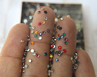 May include: Close-up of three fingers covered in colorful rhinestones. The rhinestones are various shapes and sizes, including round, square, and star-shaped. The rhinestones are a mix of colors, including red, blue, green, and orange.