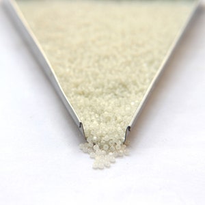 May include: A close-up of a metal tray filled with white seed beads. The beads are spilling out of the tray onto a white surface.