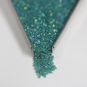 May include: A close-up of a metal funnel filled with teal iridescent seed beads. The beads are spilling out of the funnel onto a white surface.
