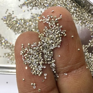 May include: A collection of numerous small, loose, faceted clear gemstones with a brilliant cut, resting on a surface. The stones are colorless and transparent, reflecting light with multiple sharp, angular facets across their surfaces.