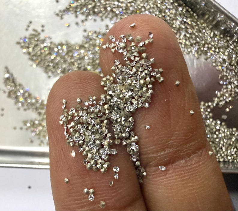 May include: A collection of numerous small, loose, faceted clear gemstones with a brilliant cut, resting on a surface. The stones are colorless and transparent, reflecting light with multiple sharp, angular facets across their surfaces.
